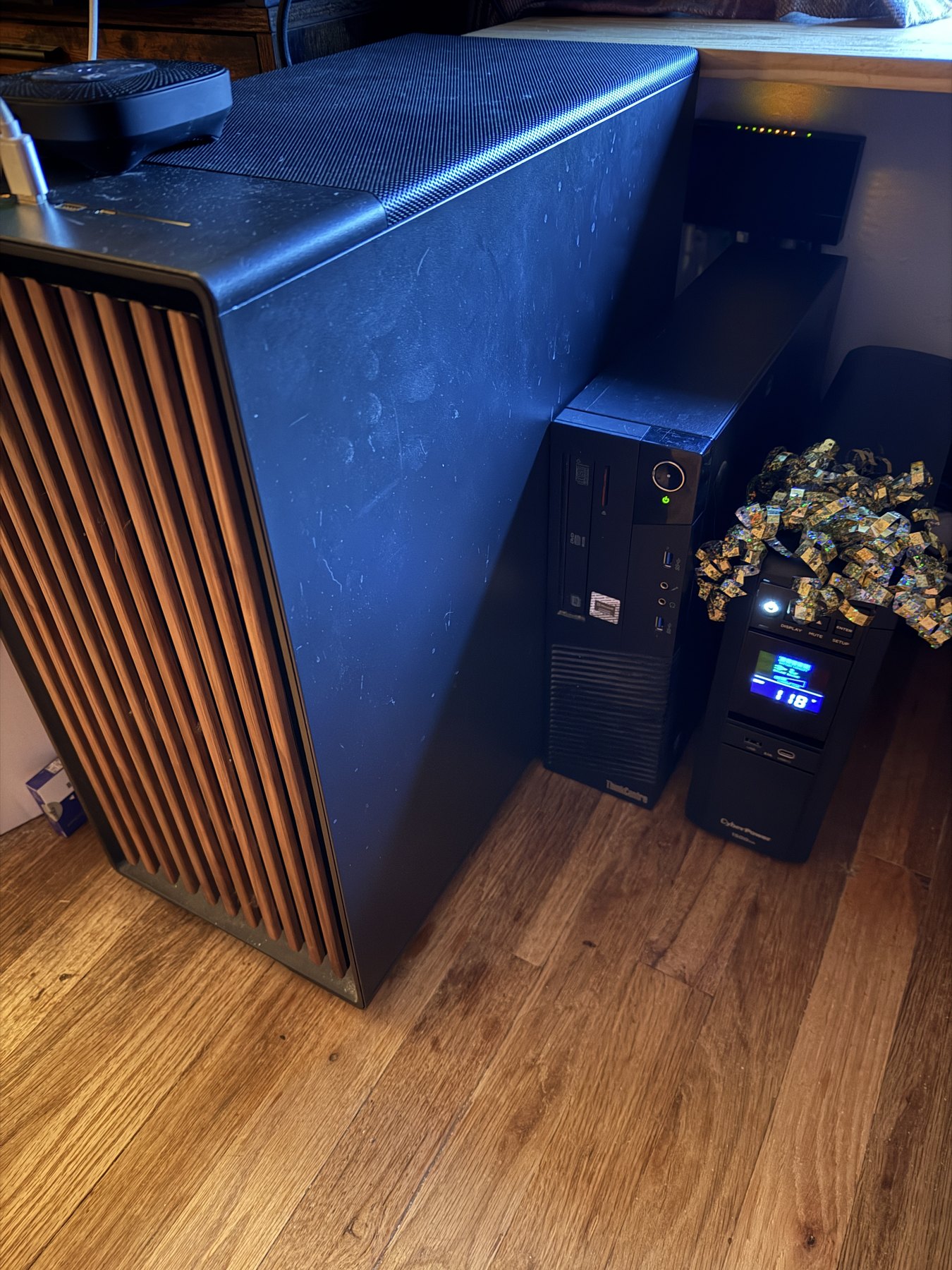 A corner of the room: a tall computer case with warm wood slats down its front, a black Lenovo ThinkCentre desktop beside it, and a CyberPower UPS with a gold holographic ribbon bow on top, its display reading 118.